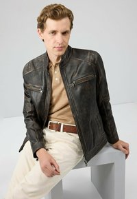 Bruine leren bomberjack met ritszakken, slim-fit ontwerp, subtiele versleten textuur, gedragen over een beige polo en crème kleurige broek.