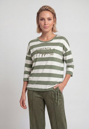 Woman with short blonde hair wearing a green and white striped shirt reading "Moments of Happiness" and green pants against a plain background.