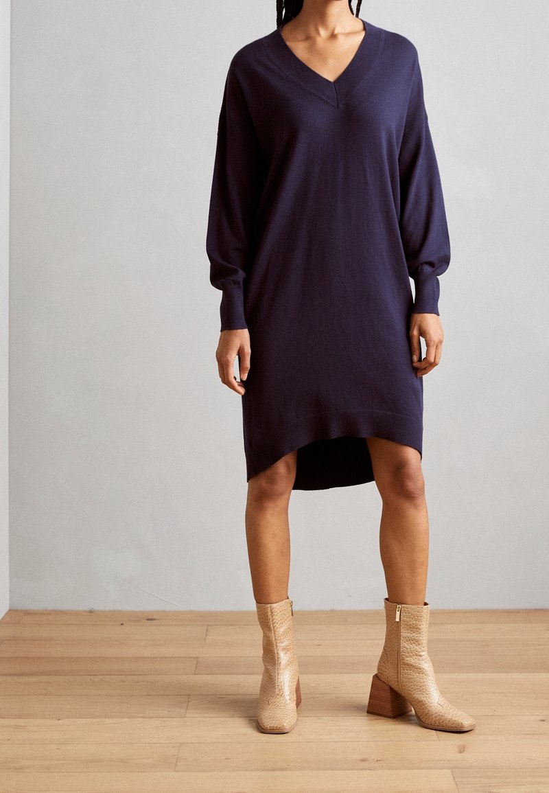 Robe tricotée bleu marine avec un décolleté en V, des manches longues et un ourlet asymétrique, associée à des bottines beige texturé avec un talon épais.