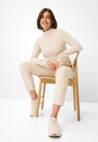 Beige lange mouwen turtleneck top gecombineerd met ribbelige beige broeken en lichte sneakers, zittend op een houten stoel met een effene achtergrond.