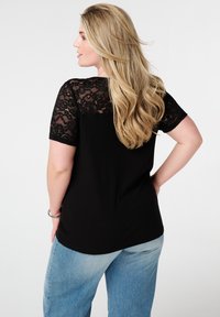 Zwarte top met kanten korte mouwen, geribbelde structuur, ronde zoom; gestyled met lichtblauwe denim jeans. Haar valt over de schouder.