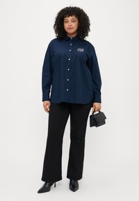 Marineblå knapp-up skjorte med spiss krage og hvite knapper, i kombinasjon med svarte flagrende bukser og en liten svart håndveske.