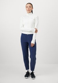 Biała sportowa kurtka na zamek z wysokim kołnierzem, w połączeniu z granatowymi spodniami dresowymi. Czarne buty do biegania dopełniają strój. Gładki, dopasowany materiał.