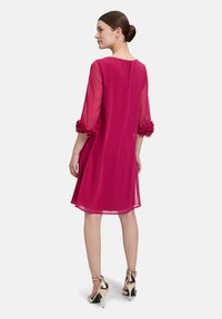 Magenta Shiftkleid mit durchsichtigen Ärmeln und Rüschensäumen. Runder Halsauschnitt und Schlüsselaktdetail im Rücken. Kombiniert mit goldenen High Heel Schuhen.