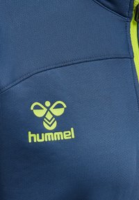 Niebieska tkanina sportowa o teksturowanym wykończeniu, z żółtym logo "hummel" i akcentami wzorniczymi, w tym kieszenią na zamek.