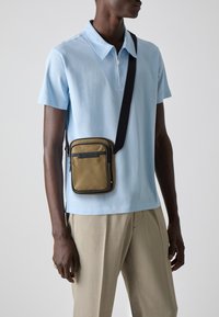 Borsa a tracolla marrone con accenti neri, forma rettangolare, texture liscia e tracolla regolabile, indossata sopra una maglietta polo azzurro chiaro.