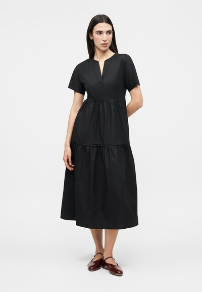 Γυναίκα με μακριά σκούρα μαλλιά, που φοράει μαύρο φόρεμα σε στρώσεις μέχρι το μέσο της γάμπας και μπορντό επίπεδα παπούτσια, στέκεται με το ένα χέρι πίσω από την πλάτη της.