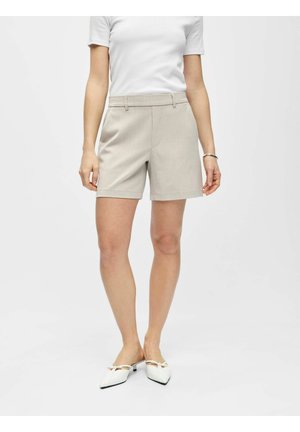 Femme portant un short beige ajusté, un top blanc à manches courtes et des chaussures blanches à talon bas et bout pointu avec des détails en nœud, debout sur un fond uni.