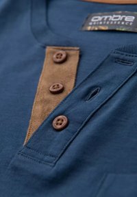 Camiseta azul marino con un tapeta de ante marrón y cuatro botones marrones. Textura de algodón suave con un pequeño detalle de lazo en el cuello.