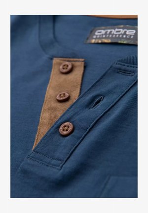 Granatowa koszulka z brązowym zamszowym detalem i czterema brązowymi guzikami. Miękka bawełniana tekstura z małą pętelką przy dekolcie.