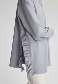 Cămașă lungă cu mâneci lungi, de culoare gri deschis, cu închidere cu fermoar și detalii cu volane pe partea laterală, fabricată dintr-un material neted și moale.