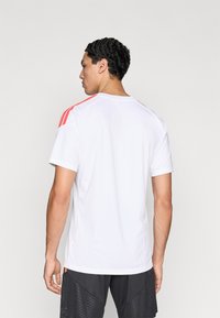 Λευκό αθλητικό πουκάμισο από απαλό ύφασμα, με κοντά μανίκια και τρεις κόκκινες ρίγες πάνω από τους ώμους. Απλός σχεδιασμός, χωρίς σχέδια.