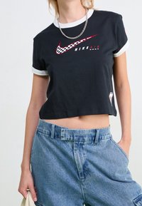 Černé zkrácené tričko s bílým žebrovaným límcem, červeným a kostkovaným logem Nike na hrudi, v kombinaci se světle modrými džínovými šortkami s vysokým pasem.