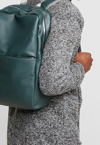 Une personne portant un pull gris à motifs porte un sac à dos en cuir vert foncé texturé avec des zips en argent visibles sur une épaule.