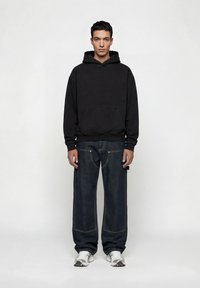 Νεαρός άνδρας που στέκεται, φοράει μαύρο φούτερ με κουκούλα, σκούρα φαρδιά τζιν και λευκά αθλητικά παπούτσια, μπροστά σε απλό φωτεινό φόντο.