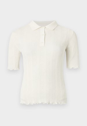 Vrouw met donker haar die een wit gebreid poloshirt met korte mouwen, donkerblauwe hoge taille wijde jeans en zwarte open hakken draagt, staand.