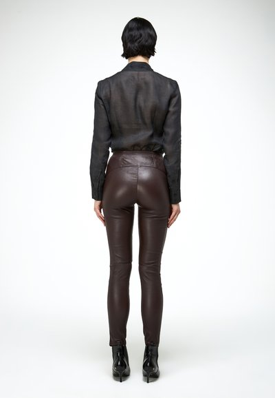 Osoba stojąca tyłem, ubrana w prześwitującą czarną bluzkę, ciasne ciemnobrązowe skórzane spodnie i czarne botki na obcasie na białym tle.