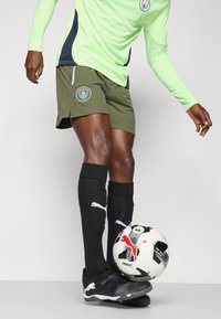 Futbolininkas, dėvintis neoninės žalios Manchester City marškinėlius, alyvinių spalvos šortus, juodas Puma kojines ir futbolo batus, valdo juodai baltą Puma futbolo kamuolį.