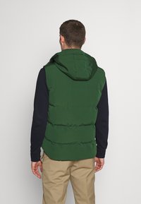 Vihreä eristetty liivi hupulla, jossa tikatut osiot, puettu tummansinisen pitkähihaisen paidan päälle, yhdistetty vaaleanruskeisiin housuihin.