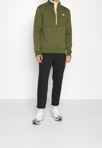 Man i olivgrön halvdragkedja Nike-tröja, svarta byxor och vit-grå sneakers står mot en helt vit bakgrund.