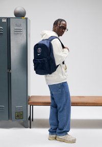 Mochila azul-marinho da Nike com o logótipo de Paris, apresenta dois bolsos frontais e alças almofadadas, usada sobre um hoodie claro e jeans azuis.