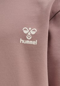 Jasnoróżowy sweatshirt z białym logo i graficznymi elementami na klatce piersiowej. Materiał wydaje się miękki i teksturowany.