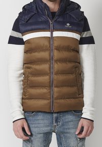 Brown puffer vest with navy hood and striped white and blue long-sleeve shirt. Features zip front, pockets, and quilted texture.