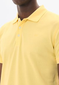 Gul polo skjorta i bomull, med krage, tre knappar och en texturerad yta. Subtil logotyp på bröstet i matchande färg.