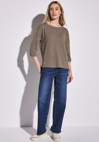 Olivově zelený pletený top s tříčtvrtečními rukávy, spárovaný s modrými širokými džíny a světle zbarvenými teniskami, na neutrálním pozadí.