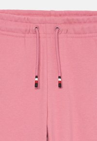 Roze trenirke s pojasom na vezanje, s krajevima kabela u crnoj, crvenoj i bijeloj boji. Tkanina je glatka i blago teksturirana.