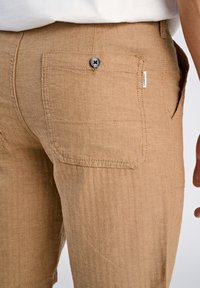 Linneblandade byxor i en ljusbrun fiskbensmönster. Har en bakficka med en svart knapp och en liten varumärkesetikett.