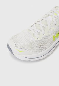 Weißer Sportschuh mit einem atmungsaktiven Mesh-Obermaterial, hellgrauen Akzenten, schwarzer Sohle und neongelber Markenkennung auf der Seite und der Zunge.