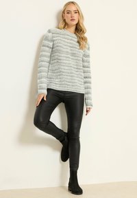 Helles graues Strickpullover mit strukturierten Streifen, kombiniert mit schwarzen Kunstlederleggings und schwarzen Ankle-Boots, vor einem neutralen Hintergrund.