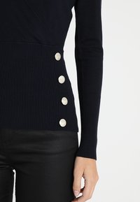 Granatowy sweter o prążkowanej fakturze, z dekoltem w kształcie litery V i czterema złotymi guzikami po lewej stronie. Długie rękawy i dopasowany krój.