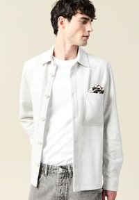 Szara koszula z guzikami, z kieszonką, noszona na białym t-shircie. Posiada gładką fakturę i klasyczny krój kołnierza. W zestawie kolorowa chusteczka do kieszeni.