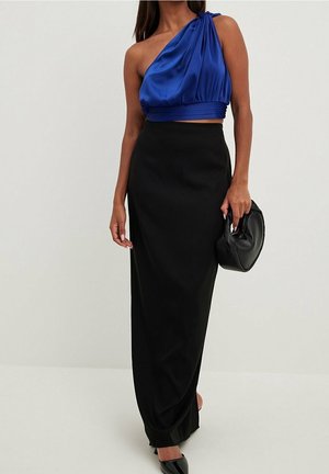 Kobieta w niebieskim topie na jedno ramię, długiej czarnej spódnicy, czarnych szpilkach, trzymająca czarną torebkę na jednolitym tle.