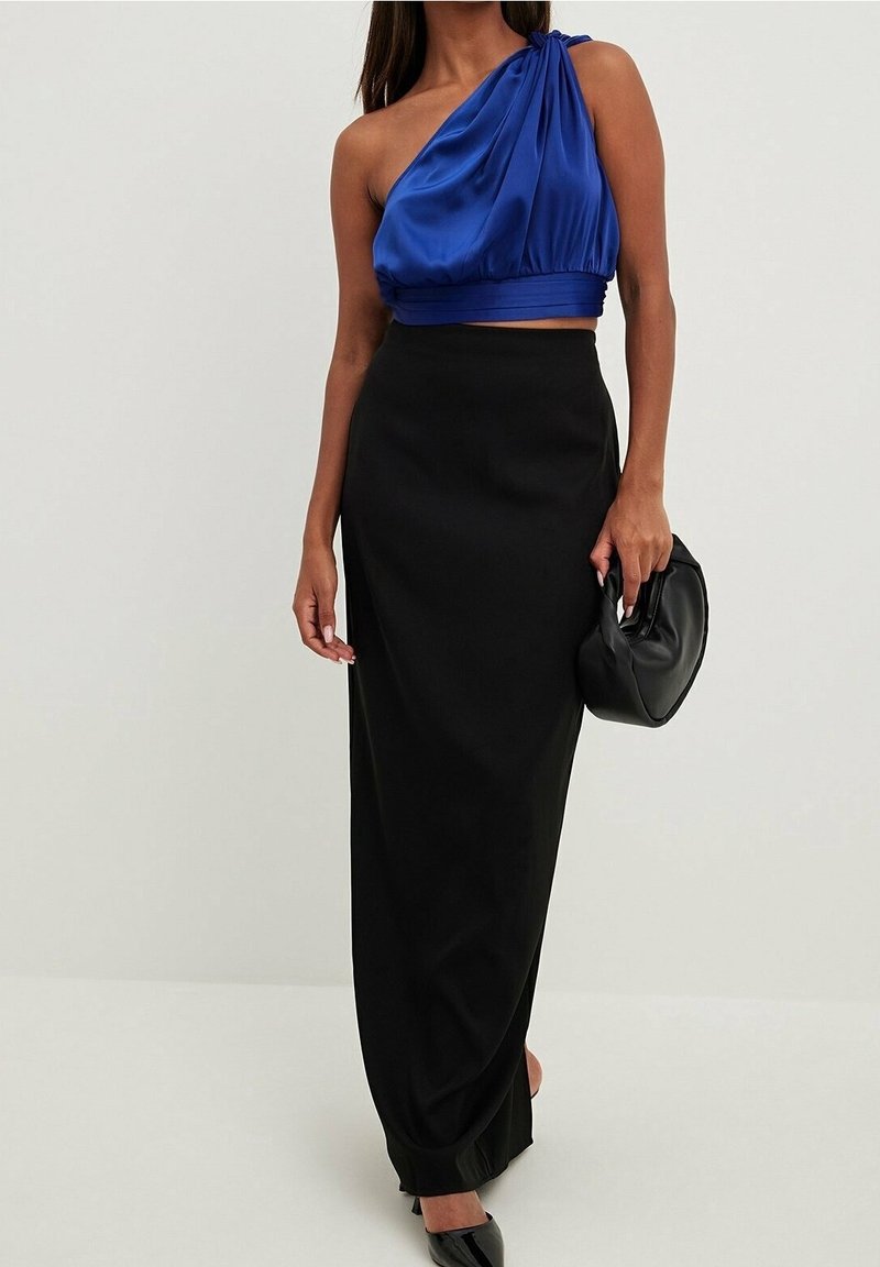 Femme portant un crop top bleu asymétrique, une longue jupe noire, des talons noirs, et tenant un sac à main noir sur un fond uni.