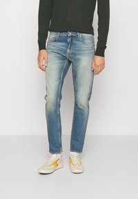 Jeans in denim azzurro chiaro con una finitura sbiadita, slim fit, cinque tasche e cuciture a contrasto, abbinati a sneakers bianche con dettagli gialli.