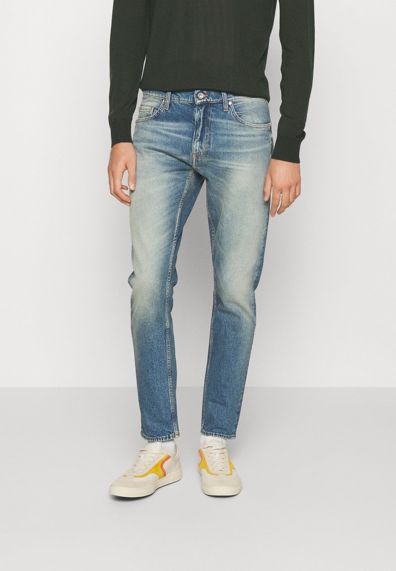Jeans in denim azzurro chiaro con una finitura sbiadita, slim fit, cinque tasche e cuciture a contrasto, abbinati a sneakers bianche con dettagli gialli.