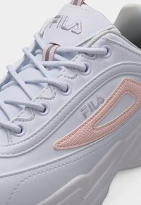 Λευκό αθλητικό παπούτσι Fila με ανάγλυφη λογότυπη ροζ ετικέτα, λευκά κορδόνια και γκρι και ροζ ριγωτή ταινία στο γλώσσα.