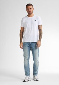Camiseta blanca con un pequeño logo, combinada con jeans de mezclilla azul claro. La persona lleva zapatillas deportivas blancas y grises.