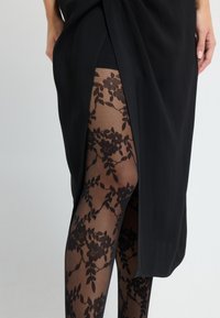 Czarna sukienka z bocznym rozcięciem, zestawiona z przezroczystymi czarnymi rajstopami z kwiatowym wzorem koronkowym, ukazującymi kontrastowy design i fakturę.