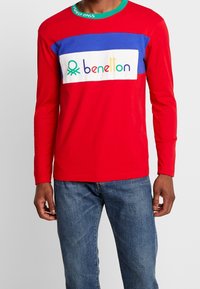 Hombre vestido con una camiseta de manga larga roja con un panel azul y un logotipo multicolor de "benetton", combinado con jeans azules sobre un fondo gris.