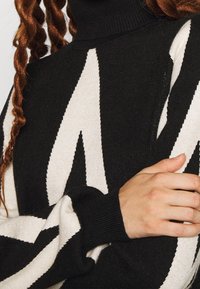 Schwarz- und cremefarbener gestreifter Rollkragenpullover mit gerippten Bündchen, der auffällige geometrische Muster aus weichem Strickmaterial aufweist.