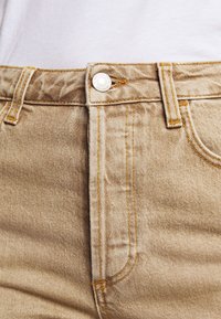 Jeans de mezclilla beige con corte recto, que cuentan con un cierre de botón plateado y costuras naranjas. La textura parece ligeramente desgastada.