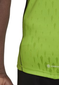 T-shirt de sport vert avec un motif texturé de formes en goutte d'eau vert foncé, doté de coutures noires et d'un logo en bas à droite.