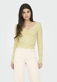 Geel lang mouwtop met een V-hals, geplooid detail in het midden, gestructureerde stof en een scalloped zoom. Gecombineerd met hooggeplaatste beige broeken.