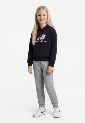 Niña joven de pie, sonriendo, vistiendo una sudadera con capucha negra de New Balance, pantalones deportivos grises y zapatillas blancas con el logotipo negro sobre un fondo blanco.