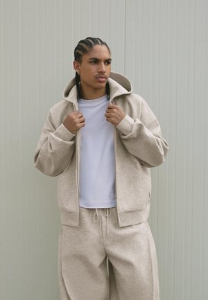 Junger Mann mit geflochtenem Haar, der einen beigen Zip-Hoodie anpasst, passend dazu trägt er eine Jogginghose und ein weißes T-Shirt, und steht vor einer neutralen Wand.