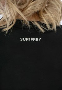 Juoda medvilnės marškinėliai su balta "SURI FREY" logo ant nugaros. Audinys yra minkštas, standartinio apvalaus kaklo dizaino.